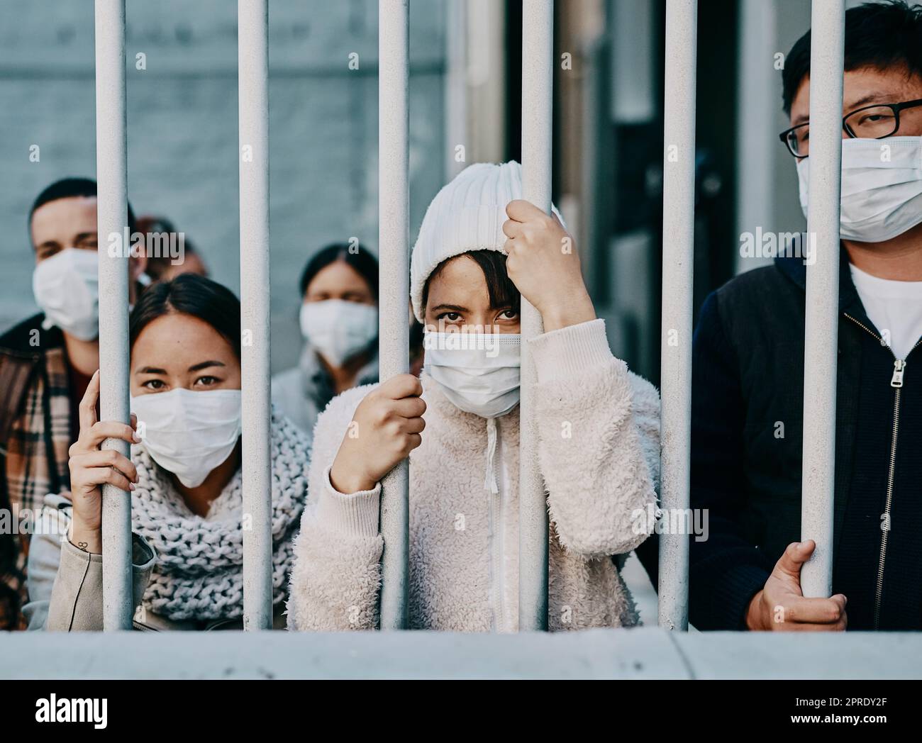Foreign people in isolation wearing covid face mask at the border or in quarantine or airport looking unhappy, upset and angry. Poor refugees, immigrants and tourists stuck behind a gate in lockdown Stock Photo