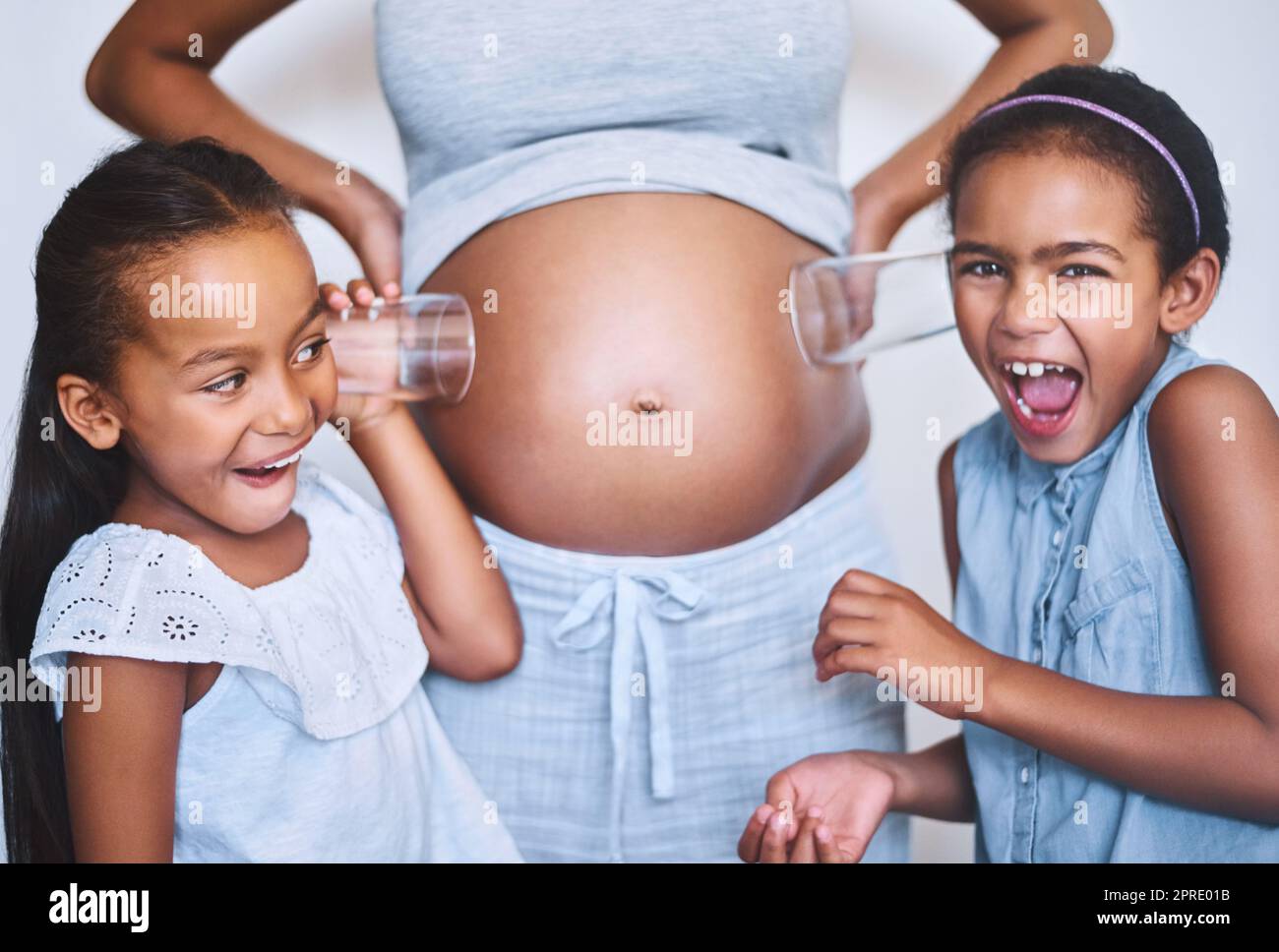 pregnant little girls I can hear him move. Portrait of two cheerful little girls standing next  their mother while each putting a glass on her pregnant belly to listen for  movement at home Stock Photo -