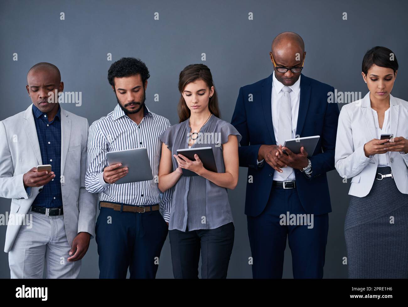 Catching up on some business while we wait. Studio shot of a group of corporate businesspeople using different devices against a gray background. Stock Photo