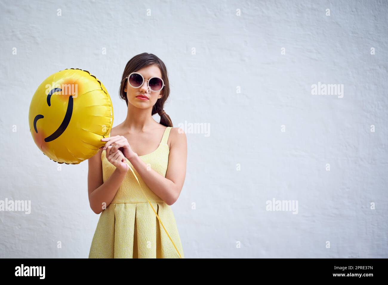 Turn that frown upside down. Studio portrait of a confident young woman holding a smiling emoticon balloon while standing against a grey background. Stock Photo