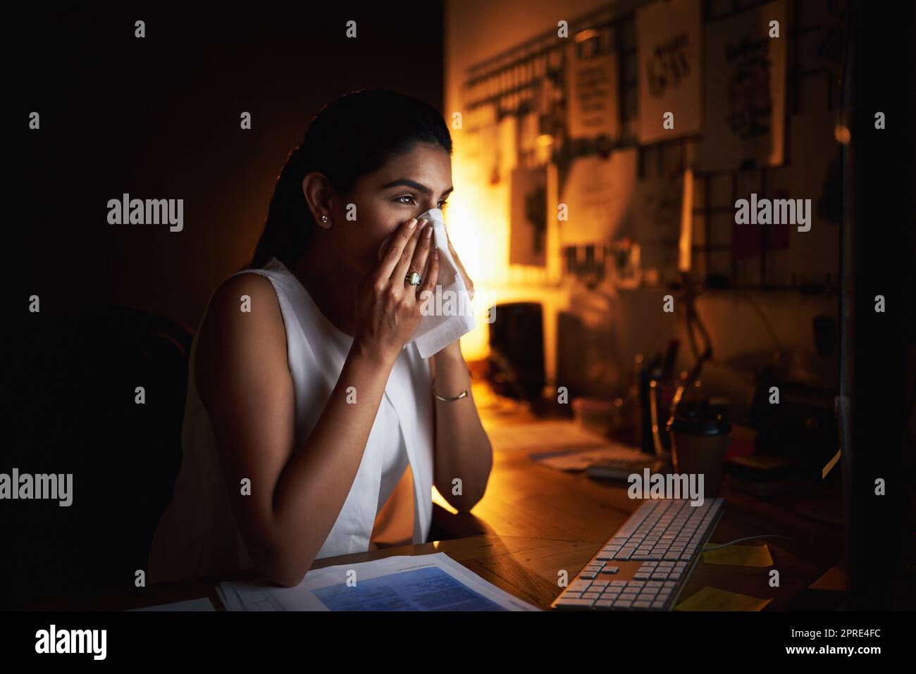 Pushing through her health concerns for the sake of success. a young businesswoman blowing her nose while working late in an office. Stock Photo