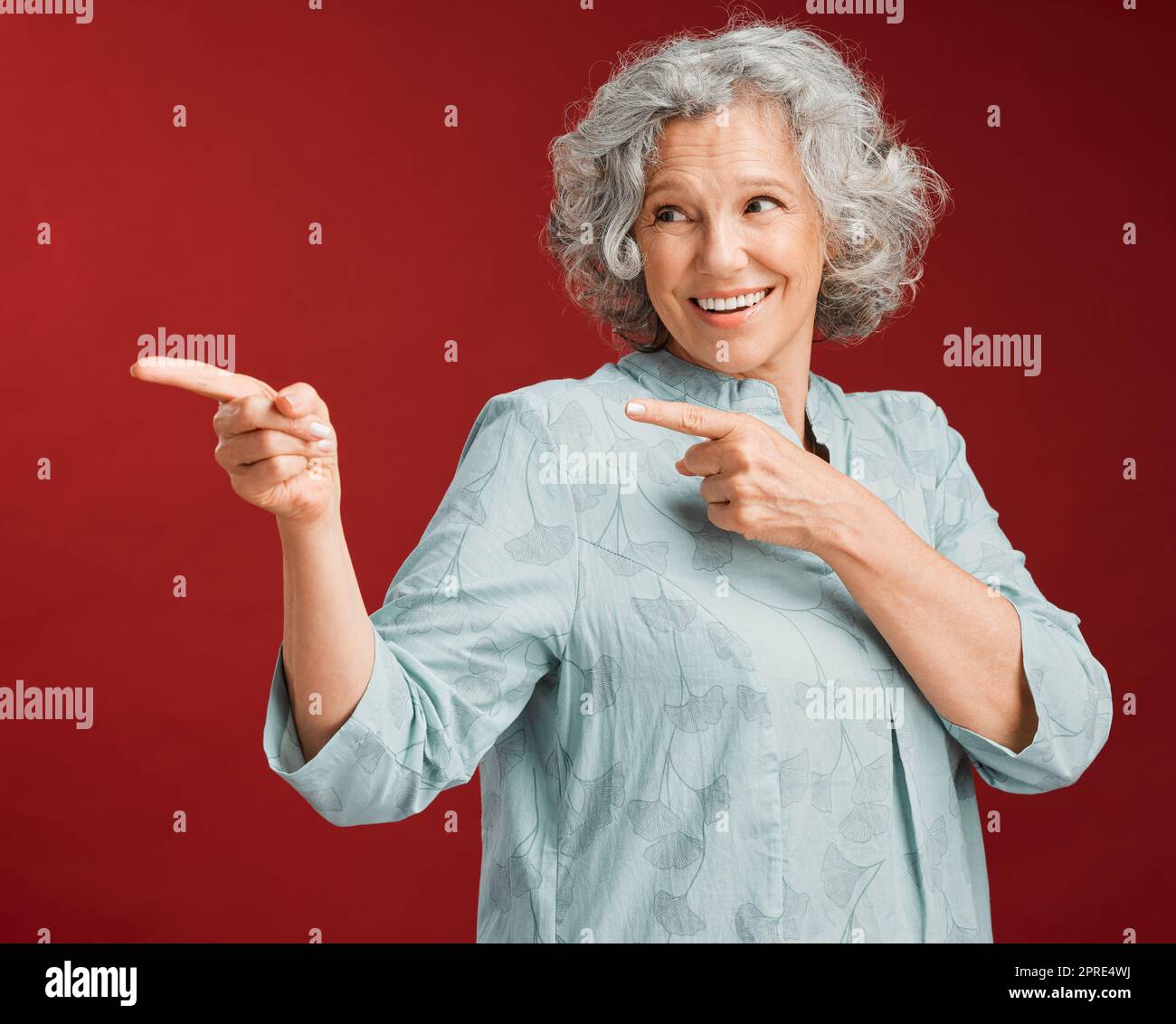 Portrait of mature woman, smiling and pointing with a gesture of affirmation, with a red background. Beautiful, confident and happy senior lady standing, index fingers pointed in positive motivation. Stock Photo