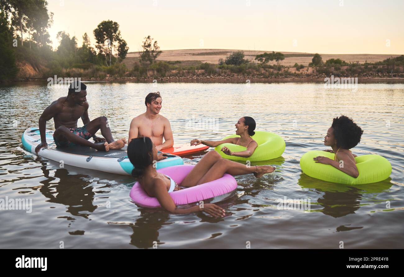 Diverse friend group floating on lake water, having fun in nature and bonding on a vacation in countryside sunset with inflatable rings. Men and women laughing, smiling and looking relaxed on holiday Stock Photo