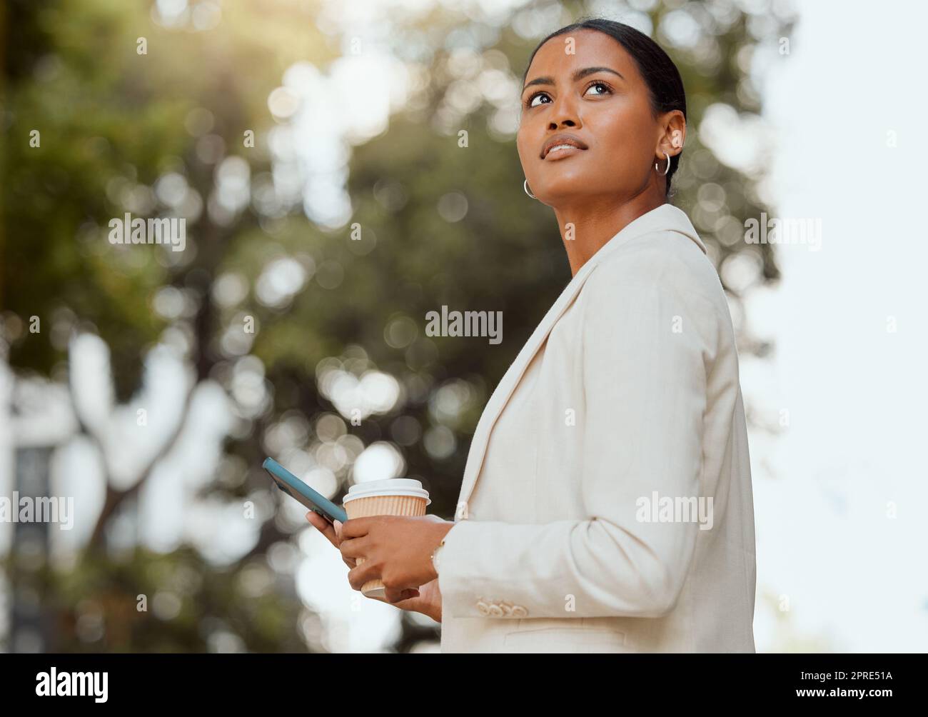 Thinking, motivation and ambition with a young business woman looking to the future with a vision and mission for growth. Female entrepreneur typing on a phone and drinking coffee outside from below Stock Photo