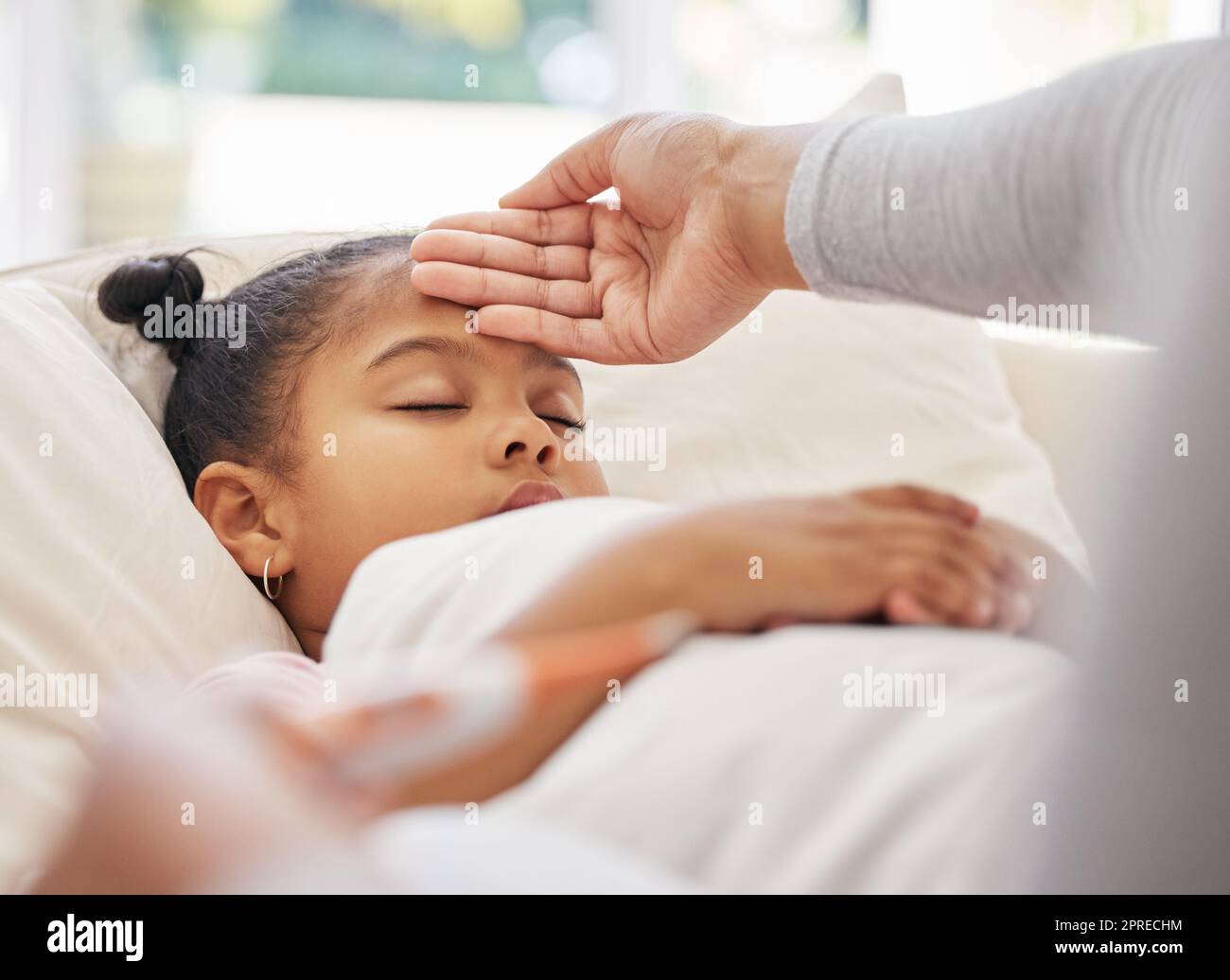 Sick little girl in bed while concerned mother uses a thermometer to check her temperature. Mixed race parent feeling daughters forehead. Hispanic chi Stock Photo