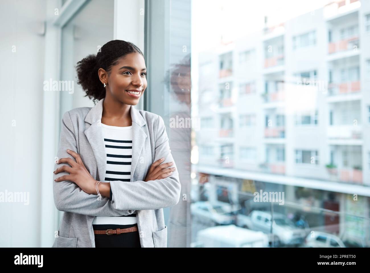 Young professional businesswoman, smiling while looking out of window of her modern office, thinking in her leadership role. Happy female manager standing with the vision of ambitious motivation Stock Photo