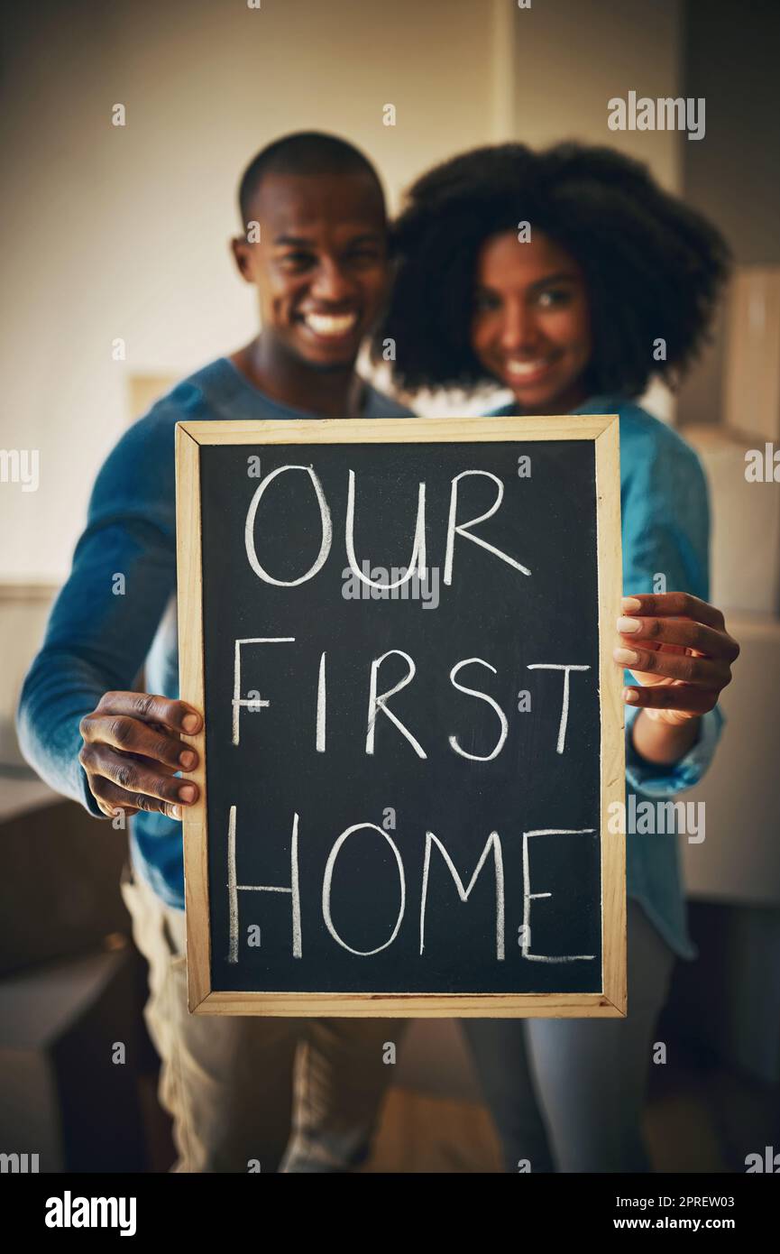 This is the new start weve been looking for. Portrait of a cheerful young couple holding up a sign saying our first home together while being surrounded by cardboard boxes inside at home. Stock Photo