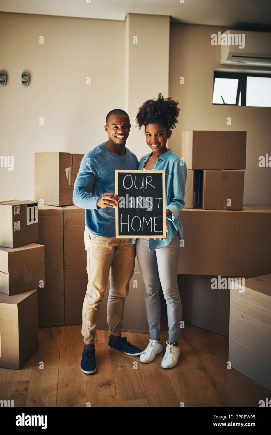 This is where our life begins. Portrait of a cheerful young couple holding up a sign saying our first home together while being surrounded by cardboard boxes inside at home. Stock Photo