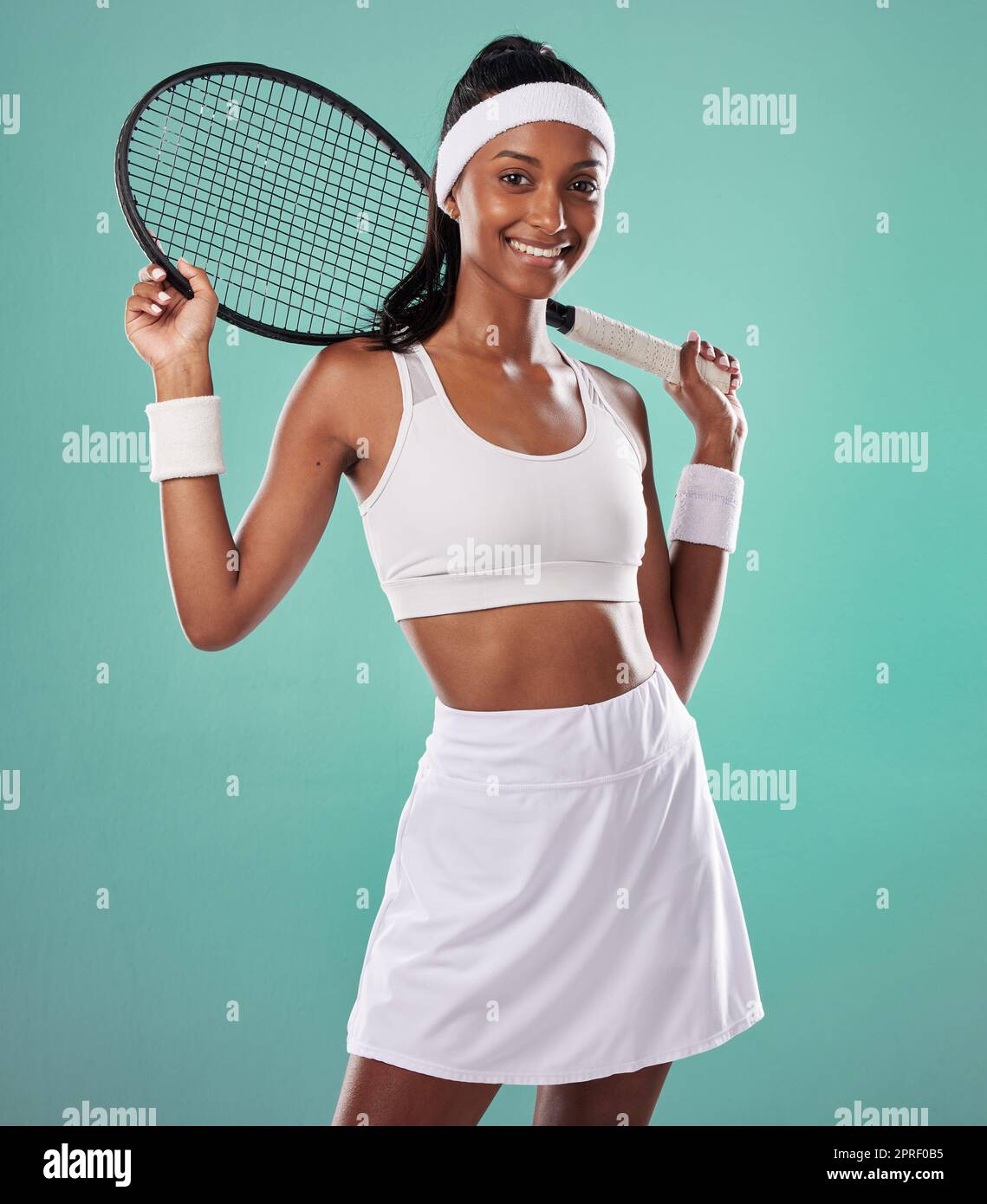 Female tennis player posing with racket, getting ready for competitive match and looking sporty while standing against blue studio background. Active, fit and happy professional sports person Stock Photo