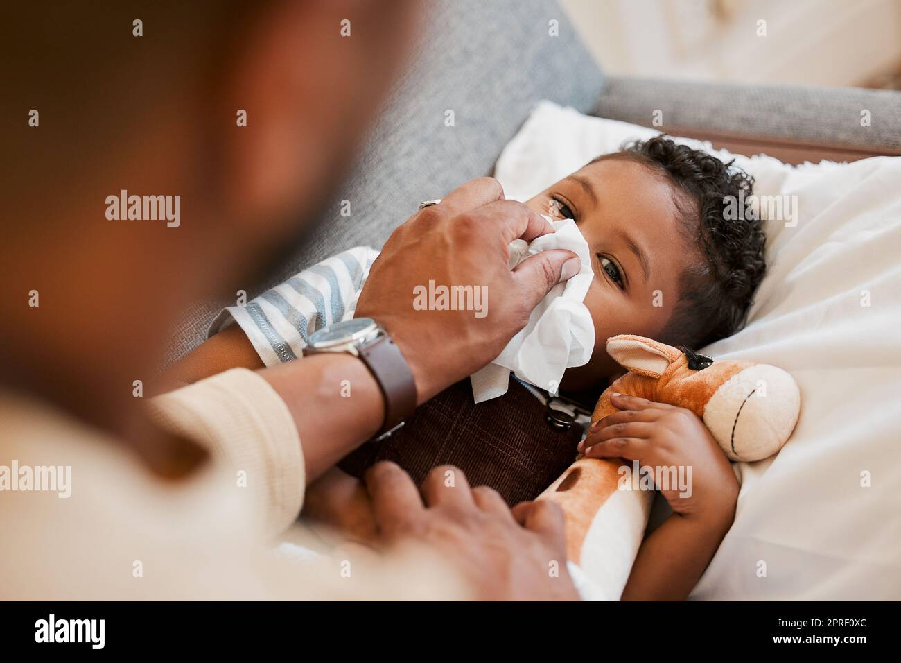 Sick, ill and unwell little boy suffering from cold, flu or covid and lying on the sofa at home while blowing his nose with dad. Cute son with a runny nose and resting in the living room from above Stock Photo