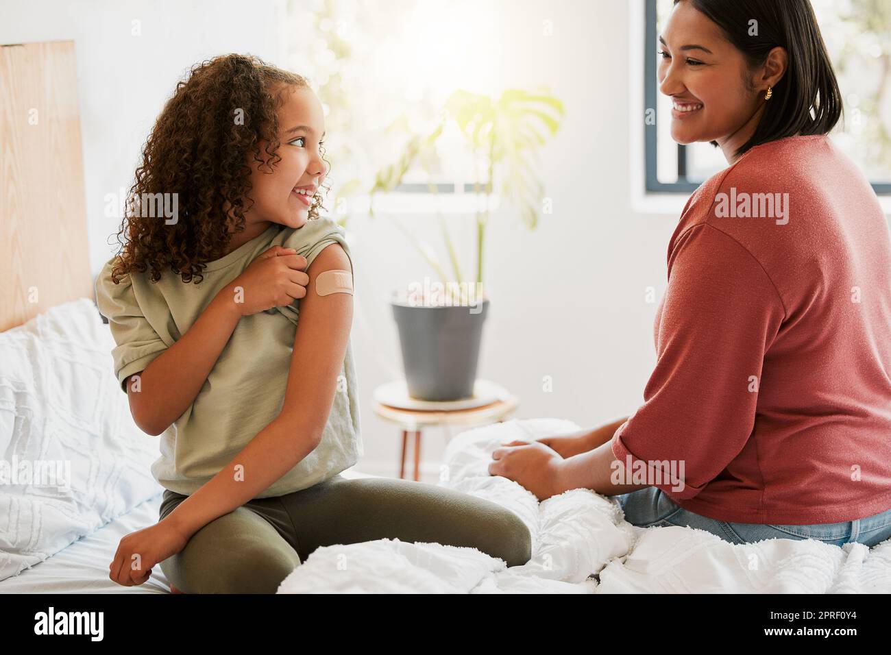 Girl with covid vaccine shows mother vaccinated arm with plaster as covid19 protection for a kid in a bedroom home. Child after getting an injection bonding with her mom in the bedroom Stock Photo
