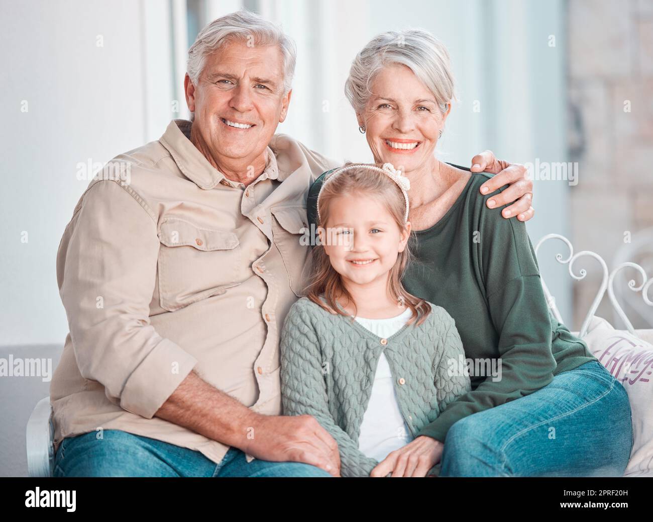 Portrait of a cheerful little girl and her grandparents sitting on the couch together during a visit at home. Loving grandparents bonding with their g Stock Photo