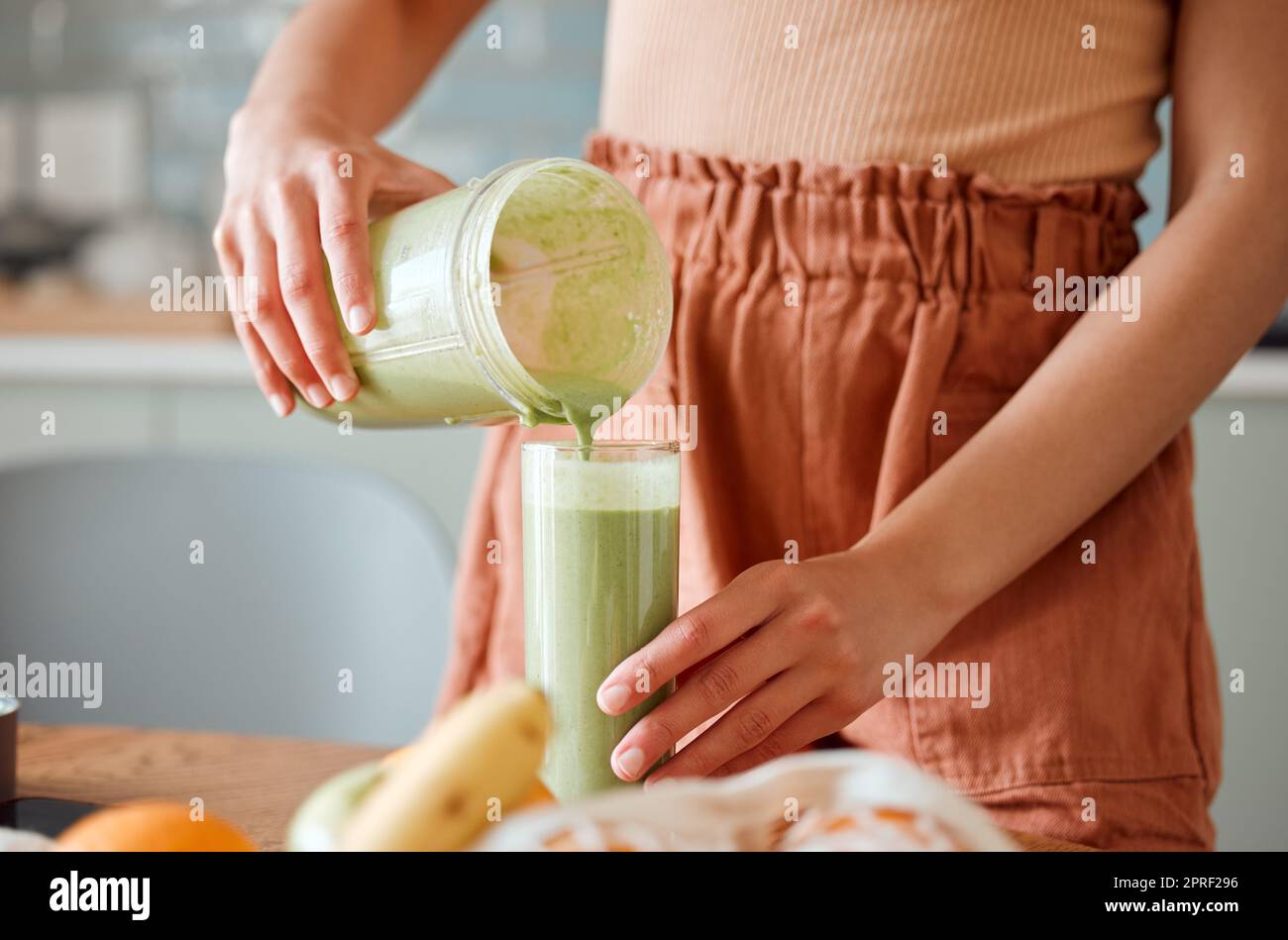 Woman pouring healthy smoothie in a glass from a blender jar on a counter for detox. Female making fresh green fruit juice in her kitchen with vegetables and consumables for a fit lifestyle. Stock Photo