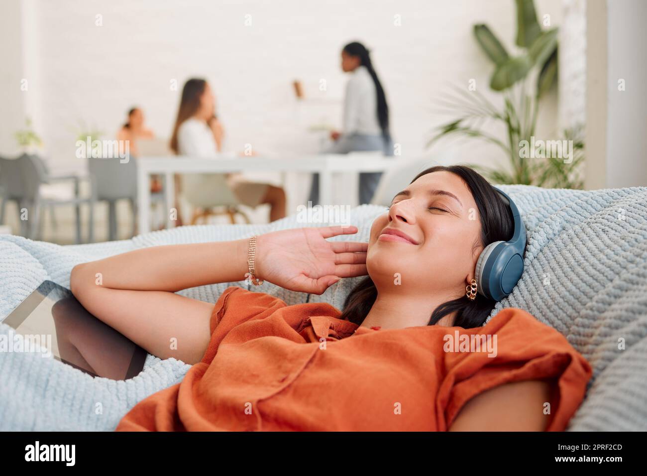 Creative wearing headphones, listening to music from tablet and relaxing, feeling inspired or motivated in a casual workspace. Happy and zen business woman lying on office bean bag and taking a break Stock Photo