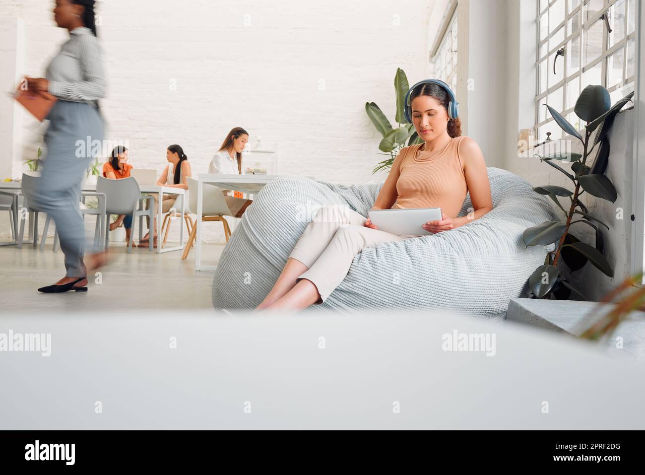 Serious, young design student relaxing and reading or listening to music in break room in startup office, with colleagues. Modern creative designer working on tablet in casual informal business Stock Photo