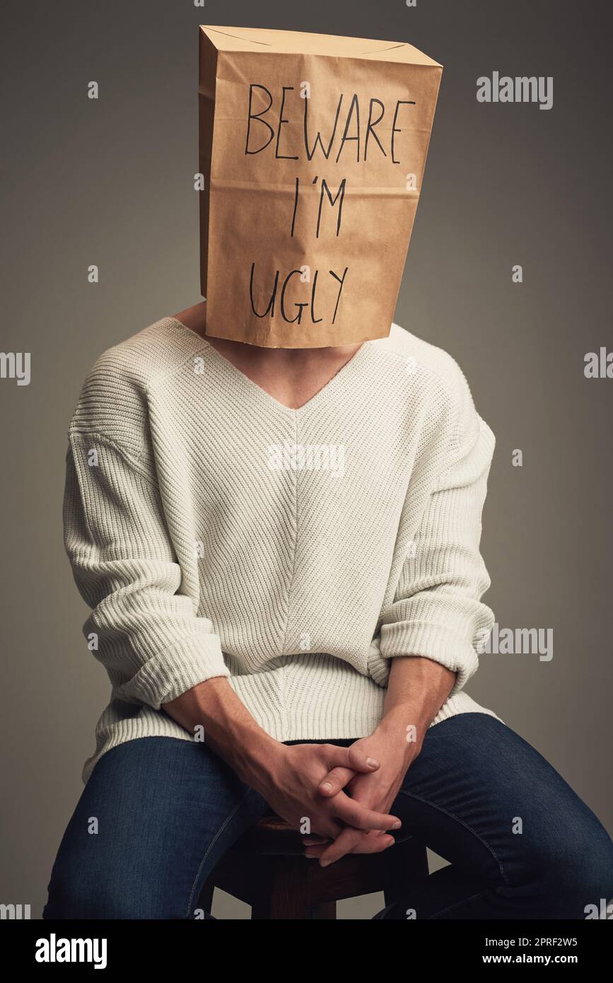 They think Im ugly but Im not. an unrecognizable man wearing a paper bag saying beware Im ugly while being seated against a grey background. Stock Photo