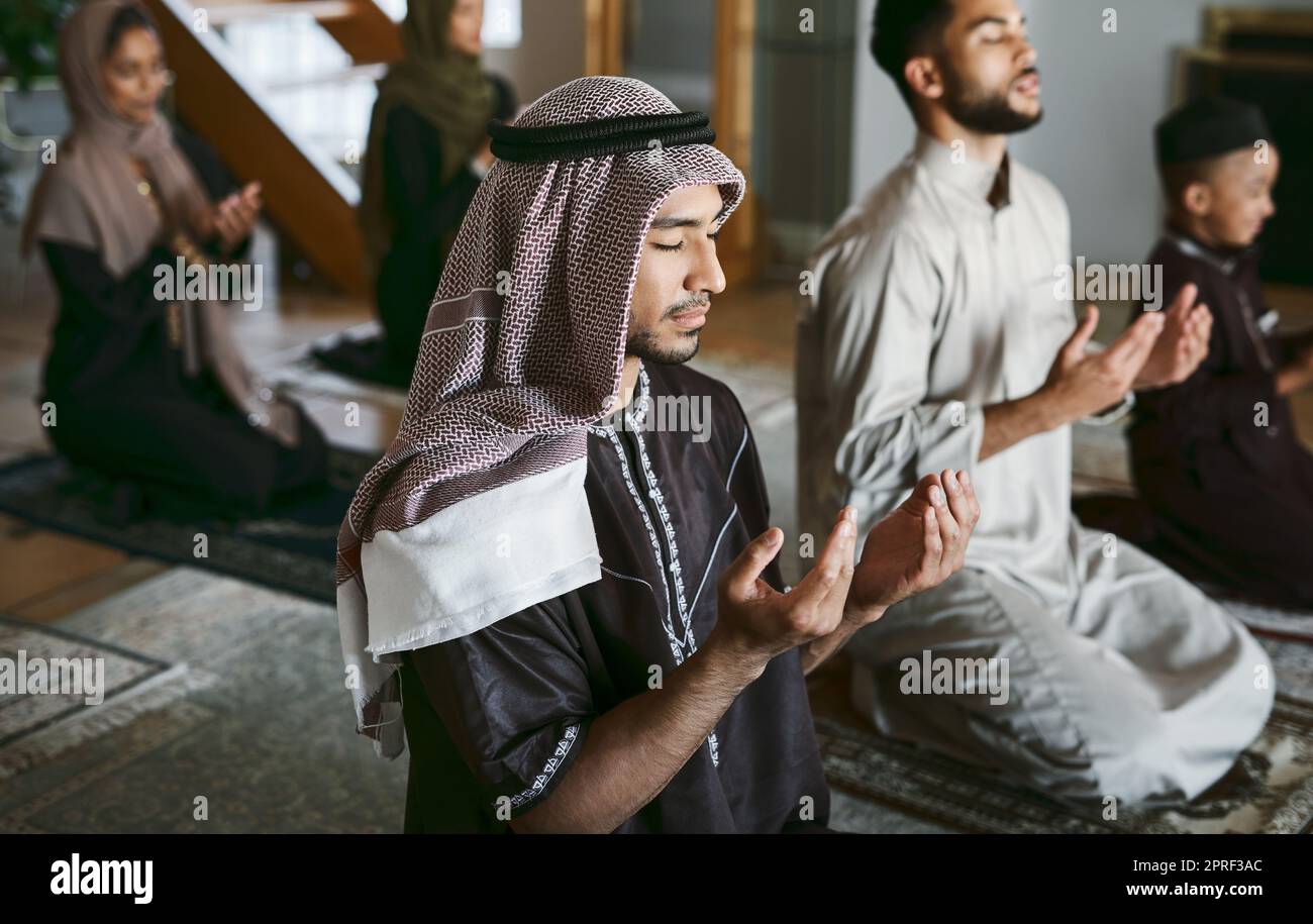 Young muslim family pray together, sit on knees at home practicing Islam. Religious women wearing hijab and spiritual men wear traditional attire, close eyes and concentrate on prayer with hands up. Stock Photo