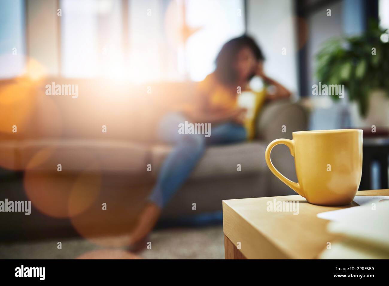 Coffee and relaxation go hand in hand. Closeup shot of a mug on a coffee table with an unrecognizable young woman relaxing in the background. Stock Photo
