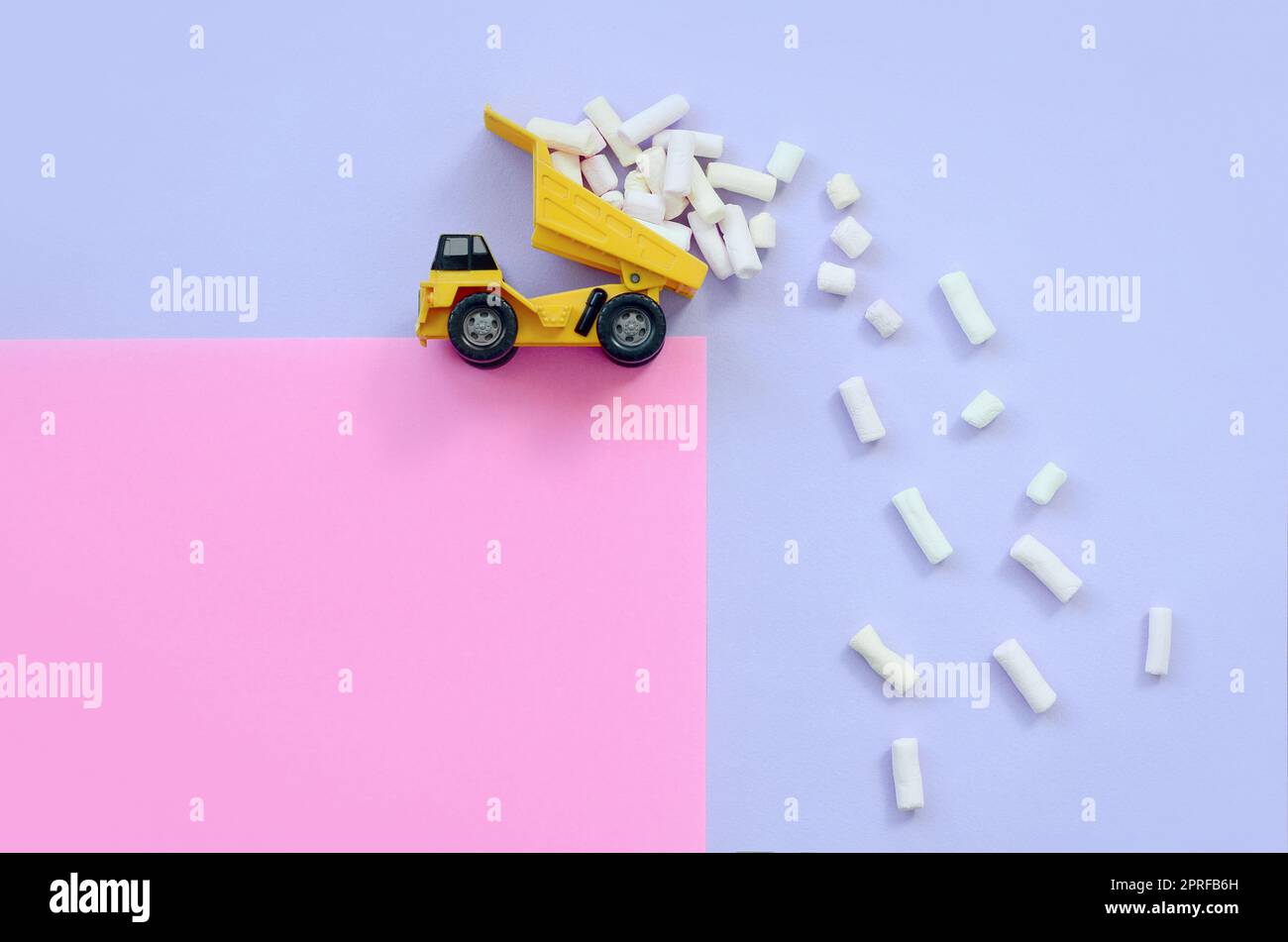 Yellow little toy dump truck throws marshmallow pieces from its raised back on a pastel violet and pink background. Flat lay minimal top view. Stock Photo