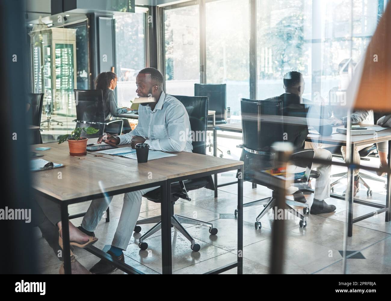 Business, office and work with a man sitting at his desk in a glass building and working with colleagues in the background. A busy corporate company with an employee and worker at a table for his job Stock Photo