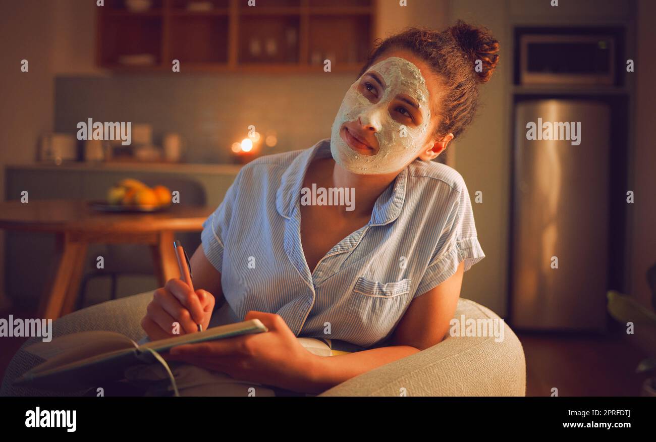 Thinking woman writing ideas in book, doing beauty face mask for skincare and facial self care sitting in living room at home. Creative female looking thoughtful while planning routine in notebook Stock Photo
