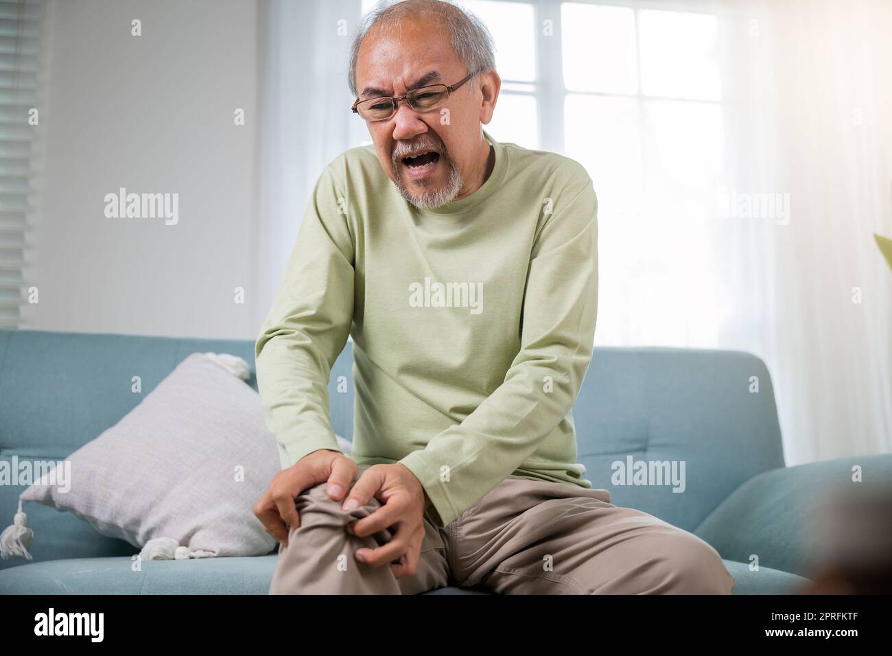 Old man suffering from knee joint pain on sofa living room, bone pain in elderly at home, senior man knee problem painful, unhappy old age hand holdin Stock Photo