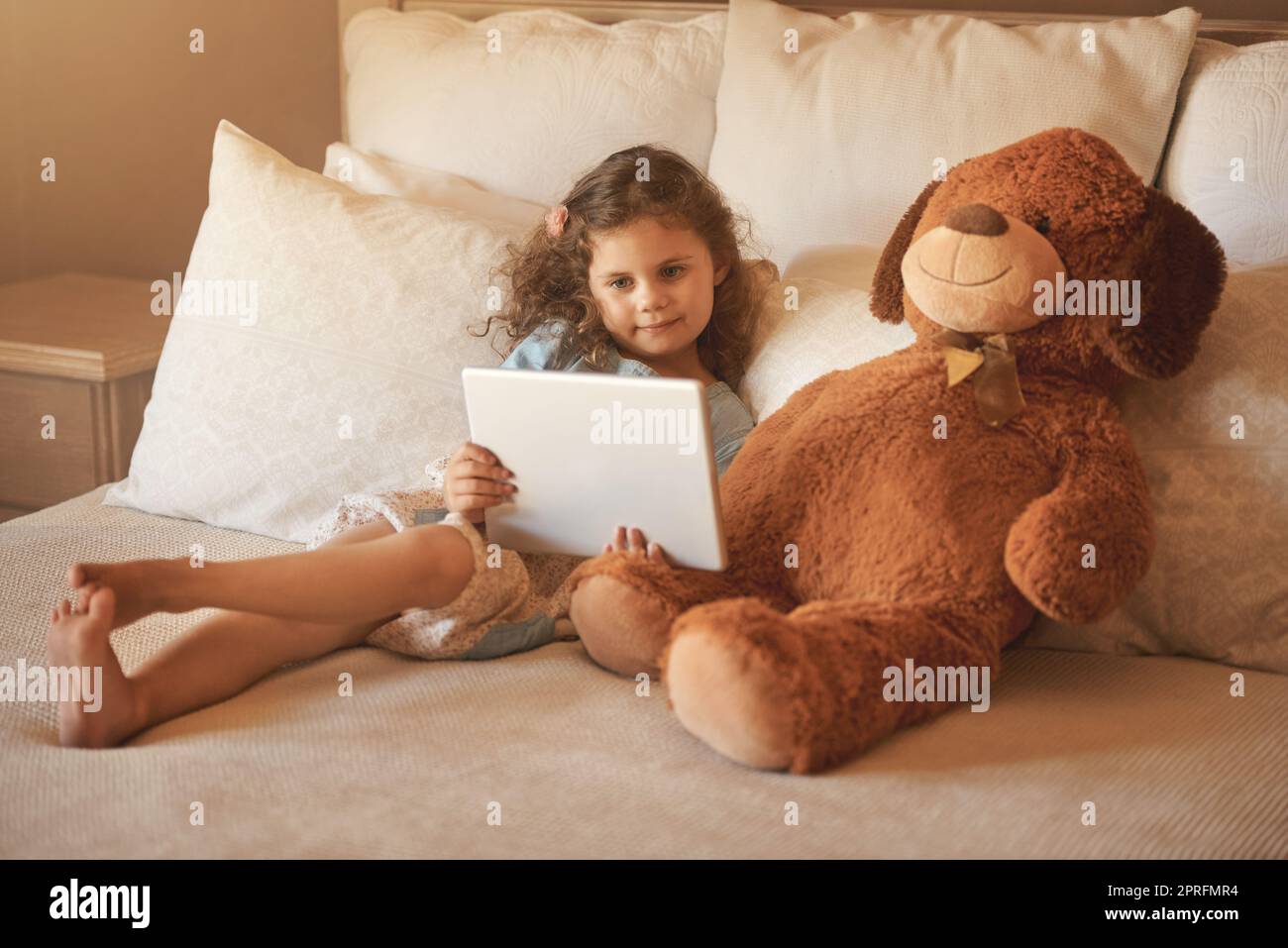 Watching a movie with my best friend. an adorable little girl at home watching a movie on a tablet with her teddybear on the bed. Stock Photo