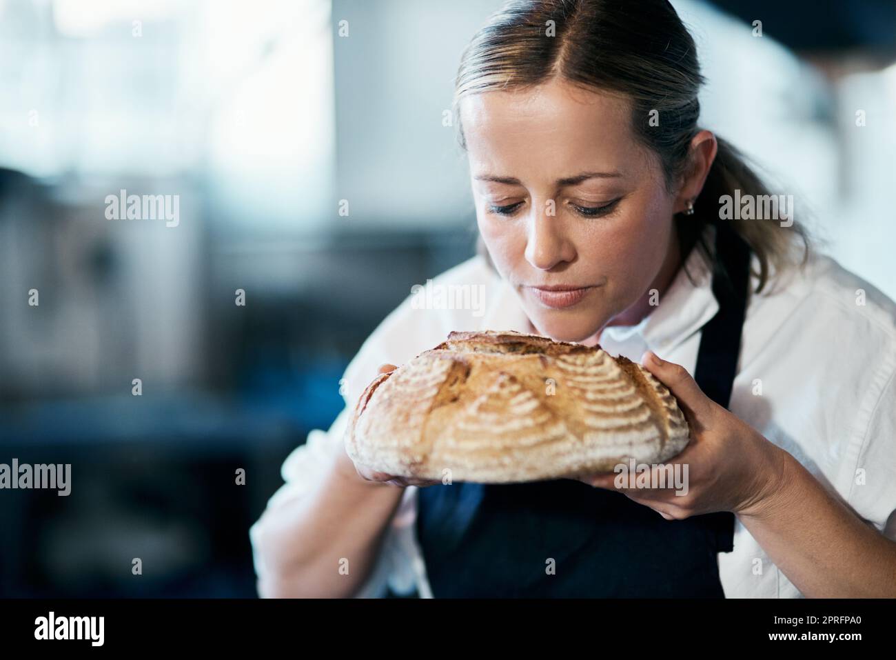 Baker, pastry chef and cafe owner smelling a loaf of fresh baked bread in the kitchen of her coffee shop. Closeup of a female cook enjoying the aroma of a freshly made dough treat or consumables Stock Photo