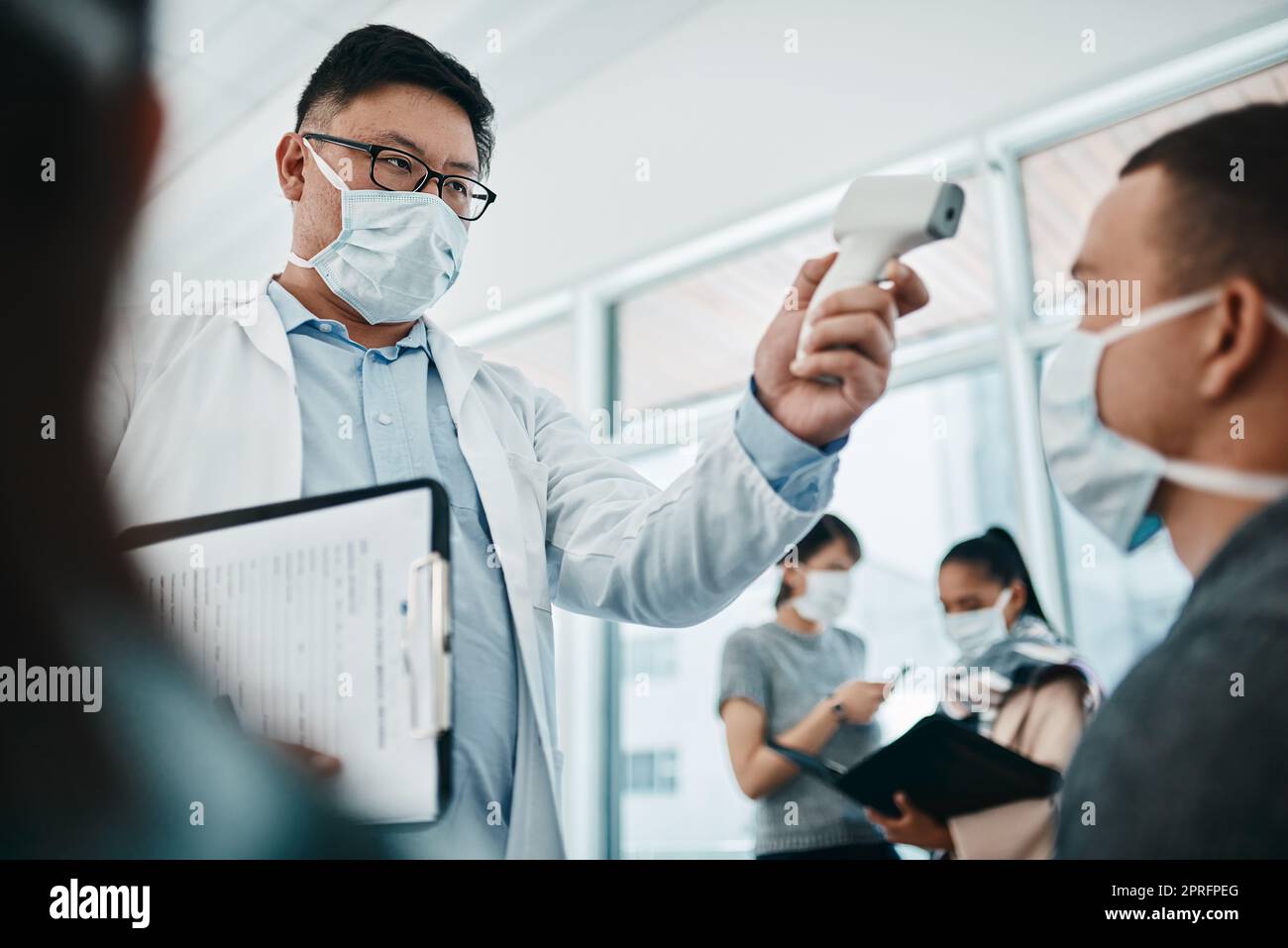 Doctor testing covid temperate in hospital appointment to prevent the spread of the virus. Healthcare compliance worker with health insurance document and face mask checking or scanning patient head Stock Photo