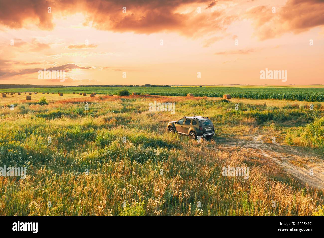Elevated View Of Car SUV Parked Near Countryside Road In Summer Field Rural Landscape. Altered Sunset Sunrise Sky Above Meadow Rural Landscape Stock Photo