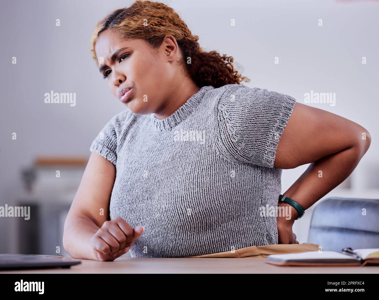 Corporate employee suffering from back pain while working at a desk in an office, uncomfortable and concerned. Young professional experience discomfort from an injury, bad posture or hurt muscle Stock Photo