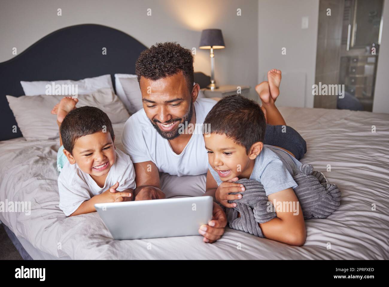 Technology, man and children relax on bed together to bond and have fun on the weekend with digital tablet. Happy father having quality time with his kids at family home for bonding relationship. Stock Photo