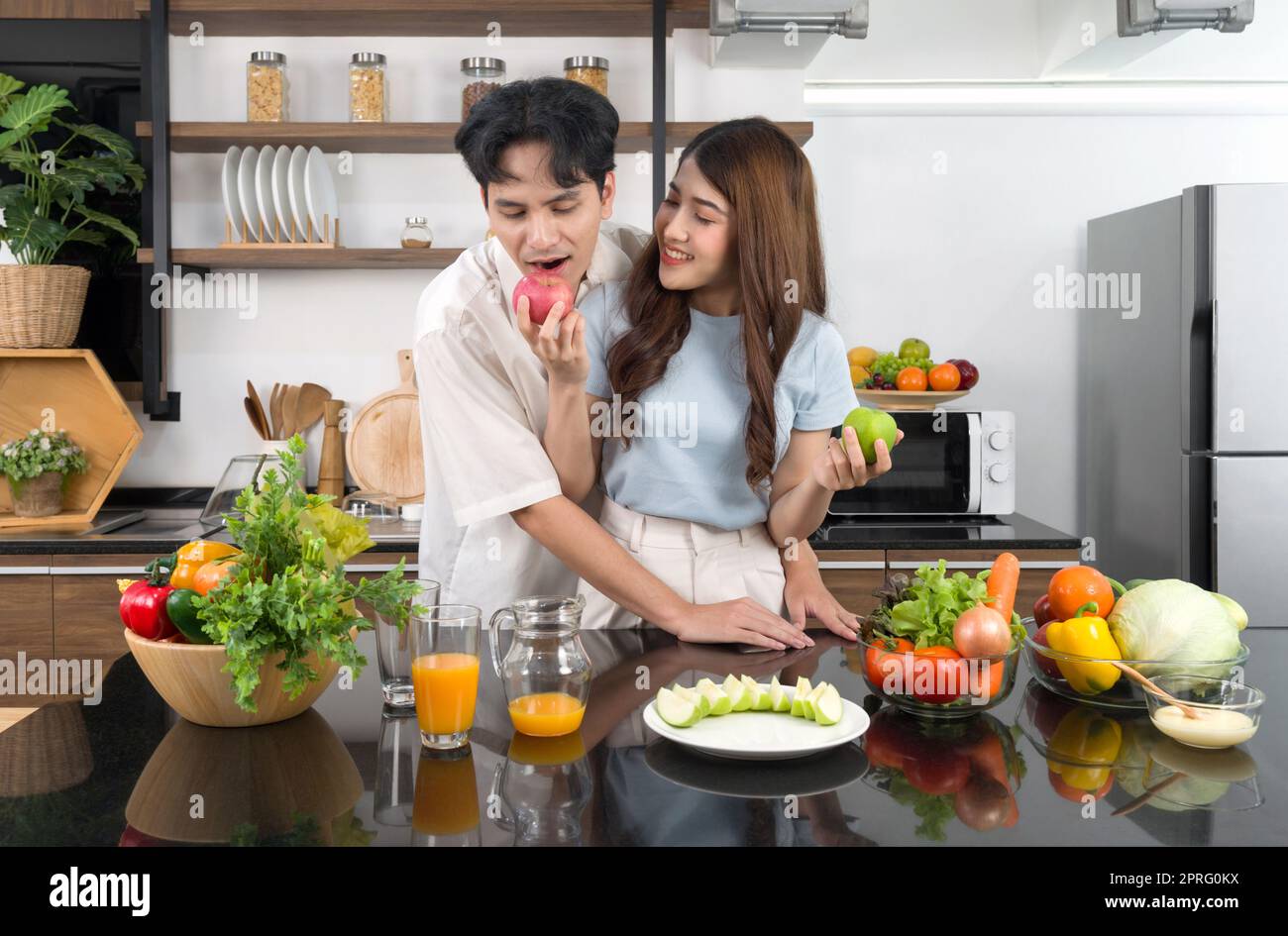 Asian couple spend time together in the kitchen. Young man in casual cloth bites the delicious red apple in his girlfriend hand. The counter full of various kinds of vegetables and fruit juice. Stock Photo