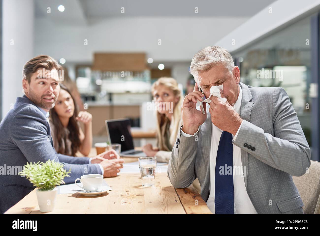 Its time to call the doctor. a businessman blowing his nose while his colleagues look on in disgust. Stock Photo