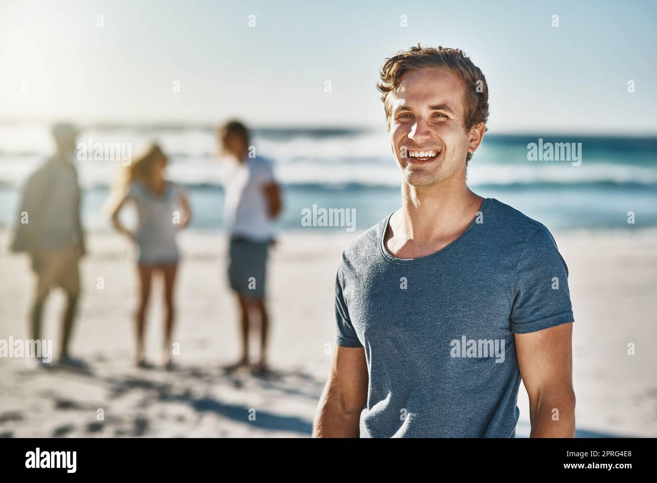 The beach is our happy place. Portrait of a happy young man posing on the beach with his friends in the background. Stock Photo