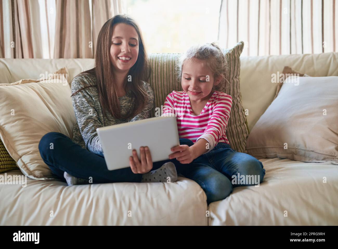 They were born tech savvy. two little girls playing with a tablet while sitting on a sofa at home. Stock Photo