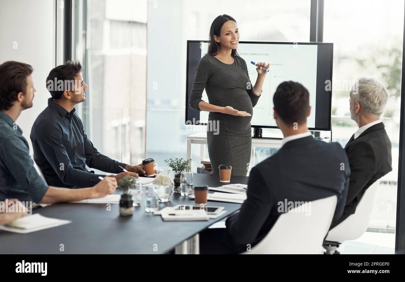 Her ideas will lead them forward. a pregnant businesswoman giving a presentation on a monitor to colleagues in an boardroom. Stock Photo