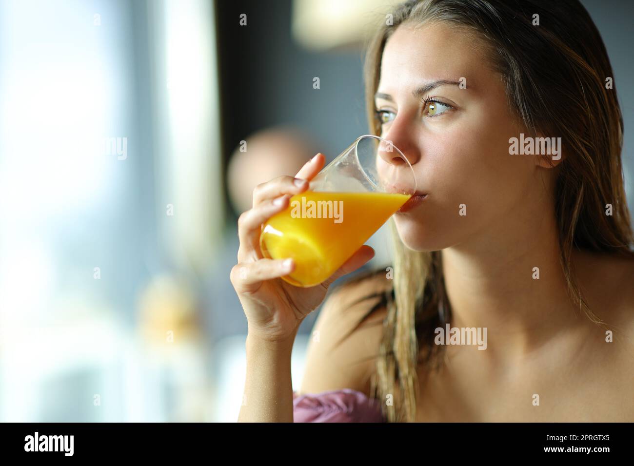 Young woman drinking orange juice looking through a window Stock Photo