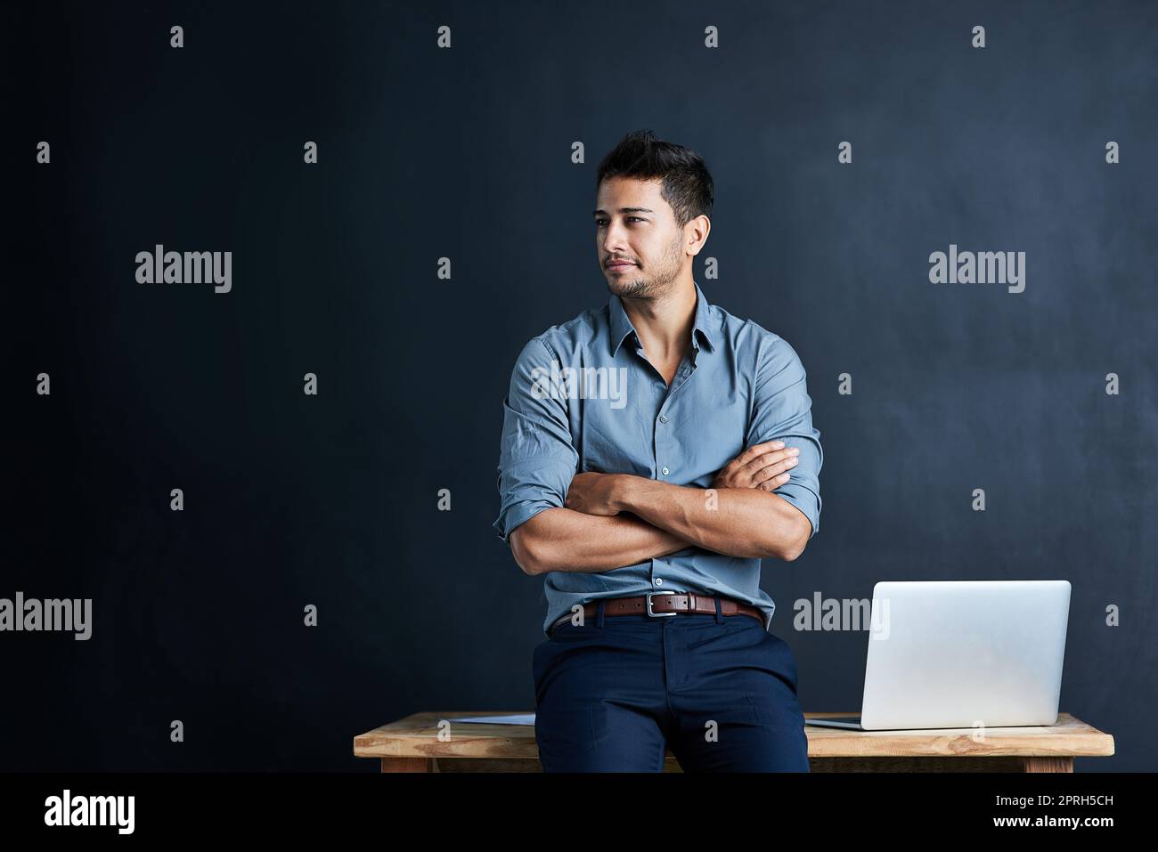 Envisioning how to start his next big success story. a handsome young businessman standing in front of a desk against a dark background. Stock Photo