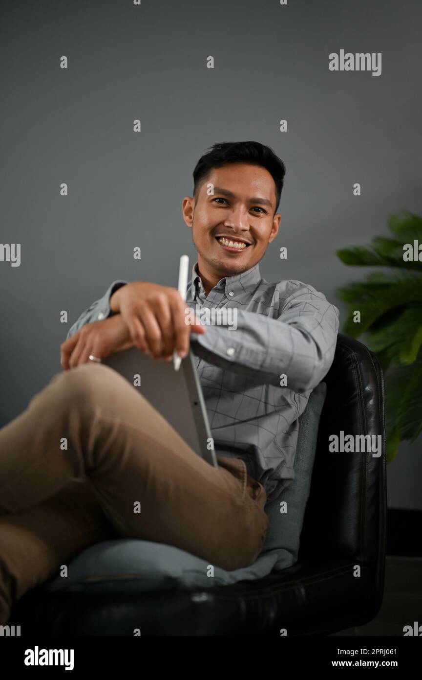 Handsome and smiling millennial Asian man in casual clothes sits on a black leather chair with his digital tablet. Stock Photo