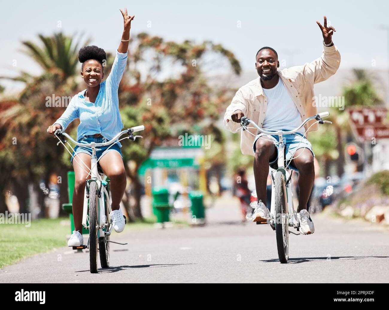 African couple cycling on bike on vacation, peace hand sign on bicycle for sustainable lifestyle in the city and happy on holiday in summer for travel. Eco friendly man and woman in Miami for spring Stock Photo