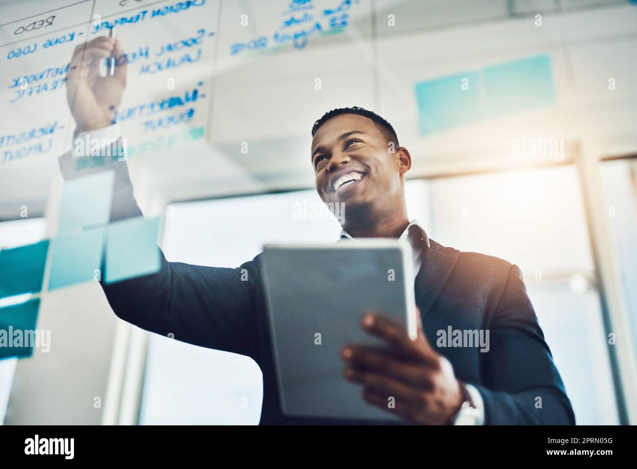 These plans are sure to revolutionize the industry. a young businessman using a digital tablet while writing notes on a glass wall in an office Stock Photo