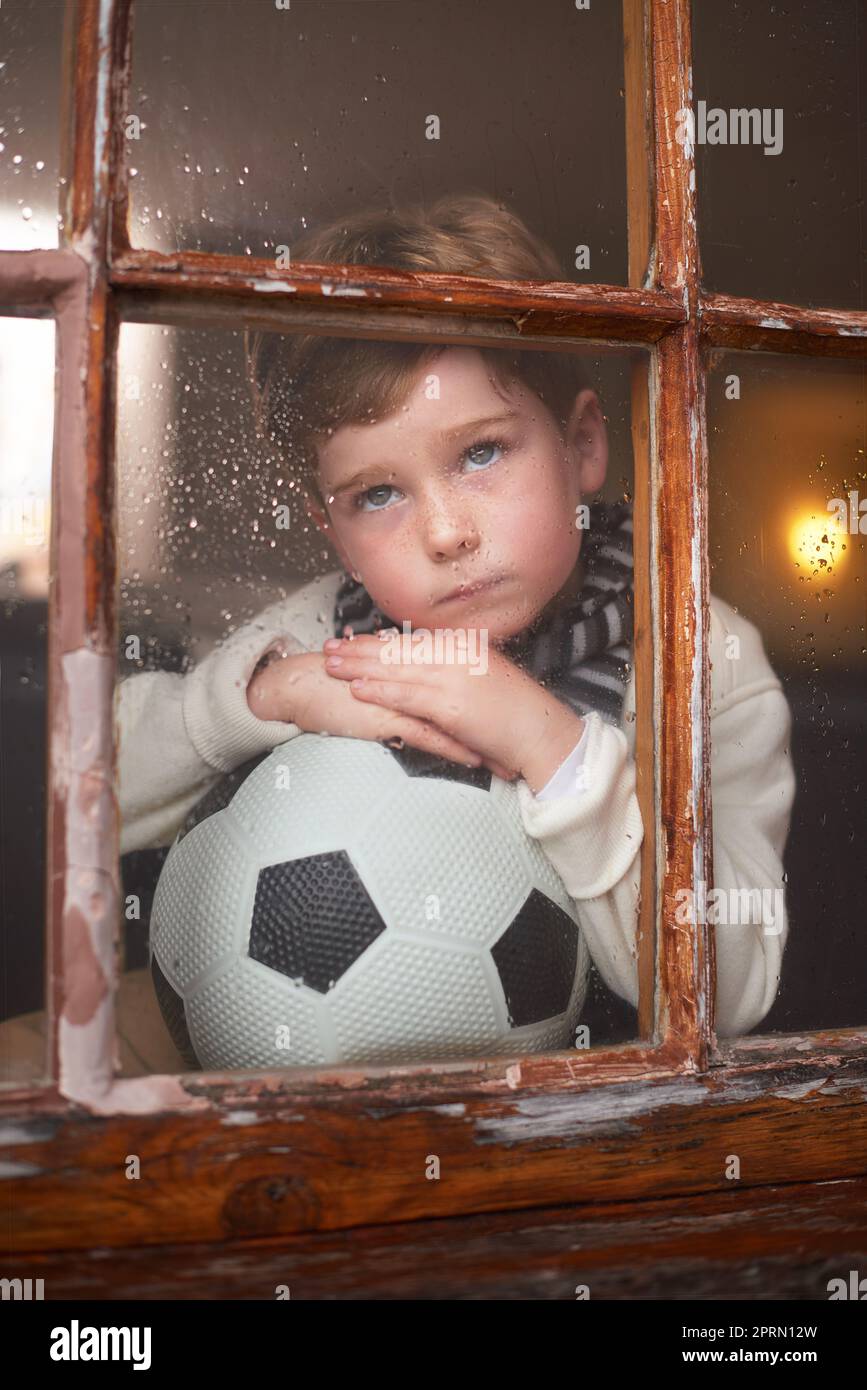 Why does it always rain when I want to play. a sad little boy holding a soccer ball while watching the rain through a window. Stock Photo