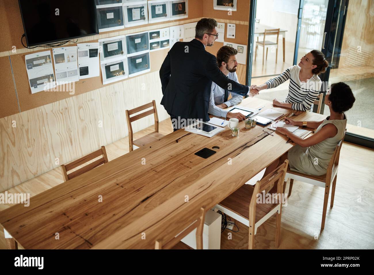 Good to have you aboard. two businesspeople shaking hands in an office while colleagues look on. Stock Photo