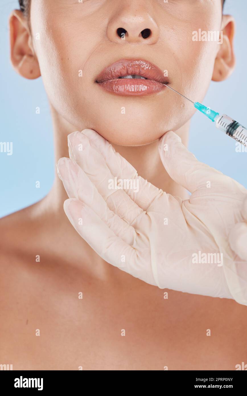 Closeup of a young woman getting lips injection treatment from a cosmetic doctor in a studio. Syringe and needle with botox filler for the mouth by a cosmetology beautician with a blue background. Stock Photo