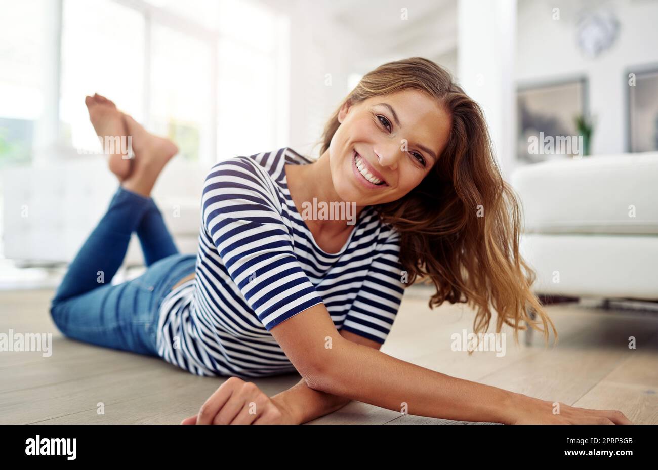 Yes my floor is so clean, I can sleep here. Full body portrait of an attractive young woman lying down on the wooden floor in her living room. Stock Photo