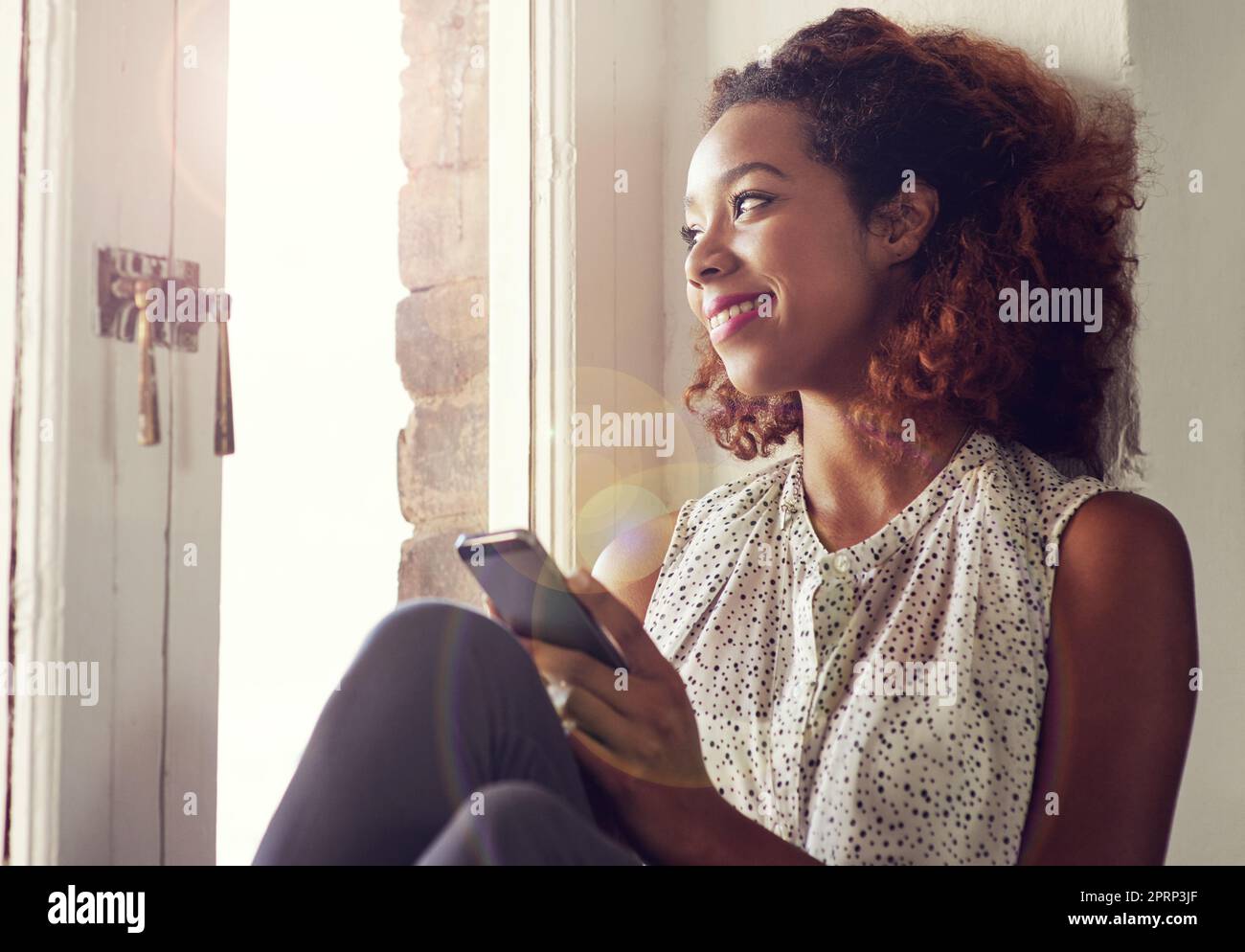 Those texts that make you feel loved...a young woman using her cellphone while sitting by a window. Stock Photo