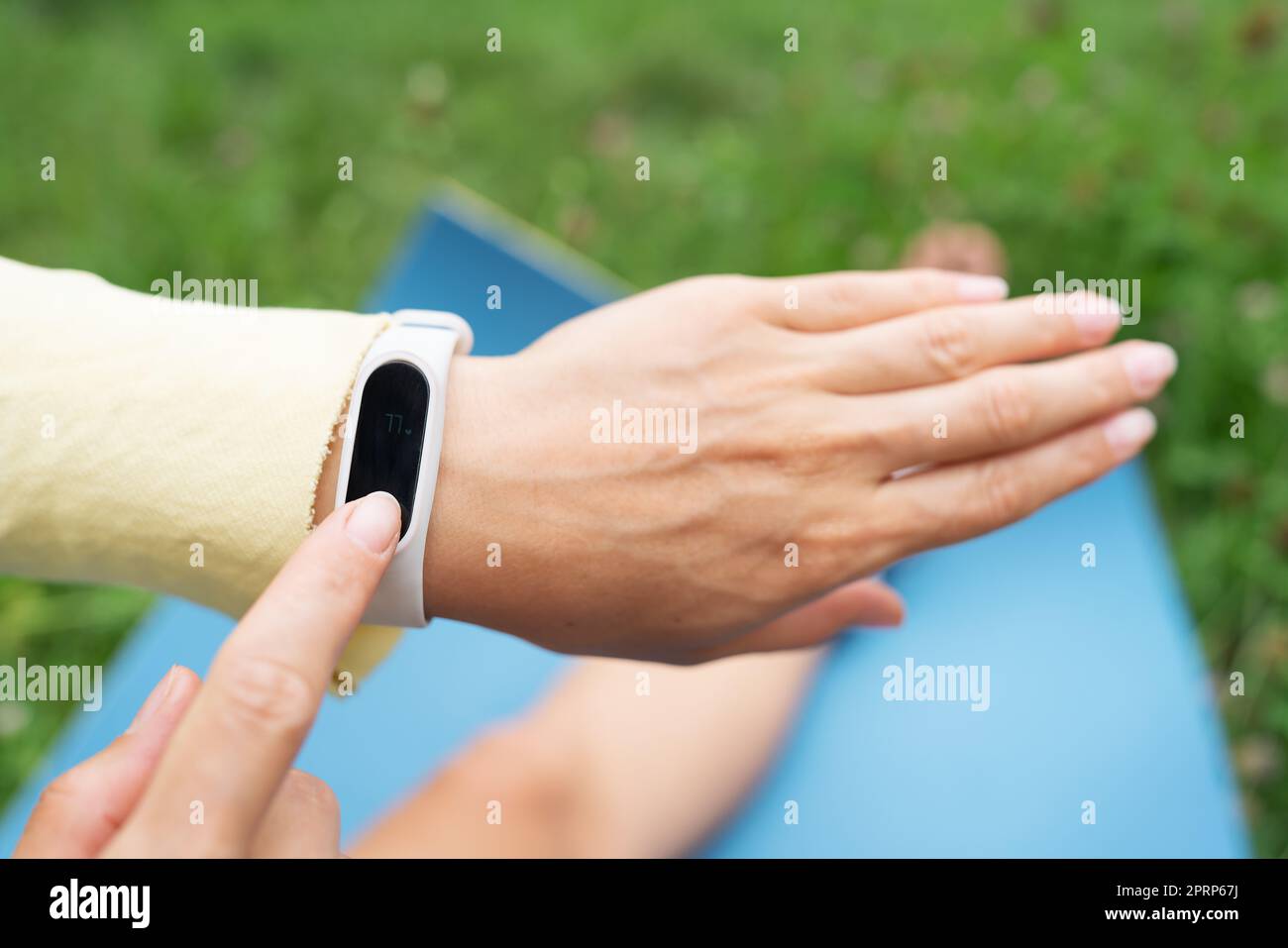 Sports girl doing stretching in nature outdoors, the girl looks at her fitness bracelet and checks her breathing during exercise. A woman is doing yoga in the park, sitting on the grass on a yoga mat. Stock Photo