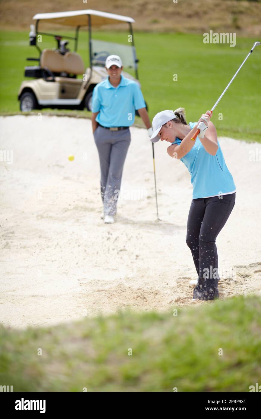 A chip off the old block. A young female golfer chipping her ball out of a bunker while her male partner looks on from behind Stock Photo
