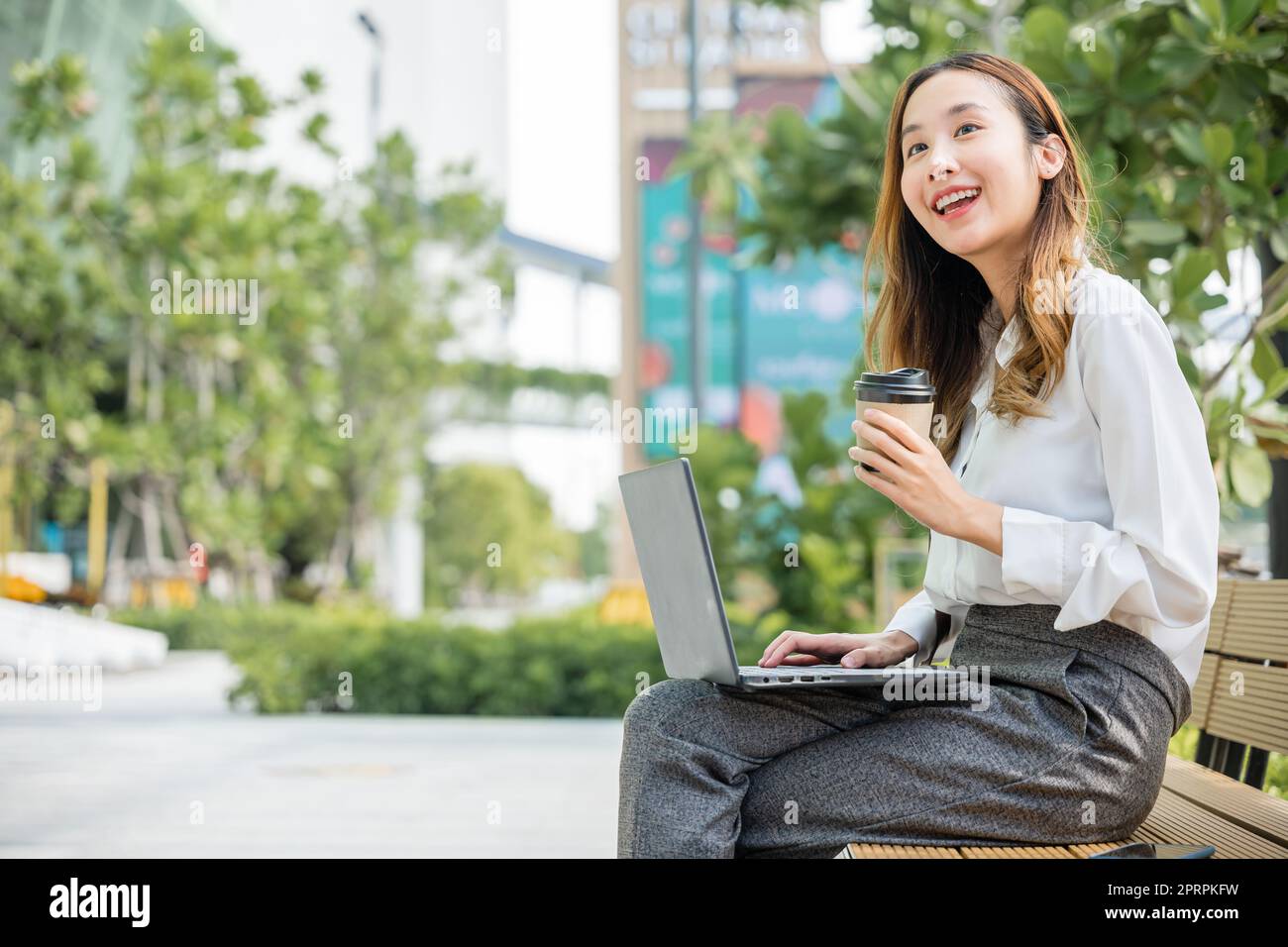 Happy Asian professional smiling businesswoman sitting alone typing computer outside street city Stock Photo
